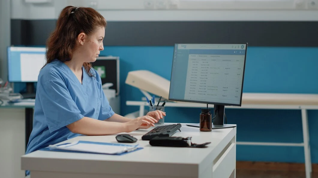 woman in scrubs sits at a desk with a computer, working on medical billing charge entry.