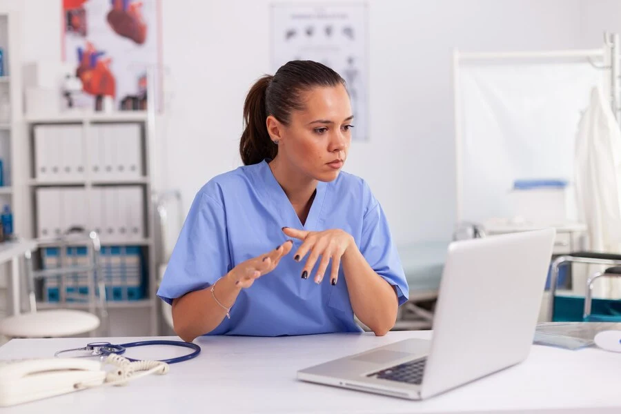 A woman in scrubs sits at a desk with a laptop, working on telehealth billing.