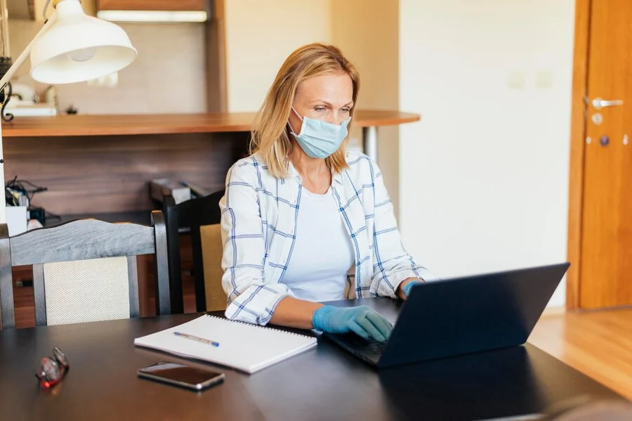 A woman in mask and gloves sits at a table with a laptop, focusing on home healthcare revenue cycle management.